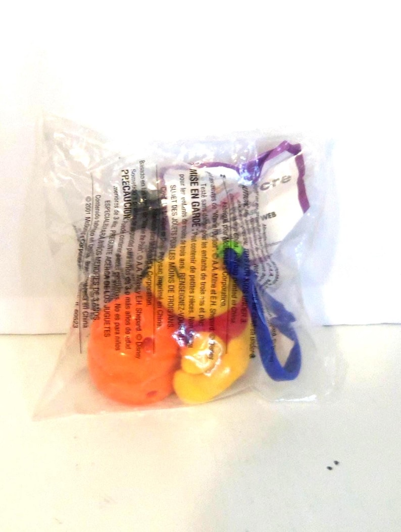 K&ouml;nnte beinhalten: Ein versiegelter, durchsichtiger Plastikbeutel mit einem kleinen Spielzeug, das eine leuchtend orangefarbene, abgerundete Basis, einen gelben Abschnitt und eine blaue Kunststoffschlaufe aufweist. Die Verpackung ist mit Sicherheitshinweisen bedruckt.