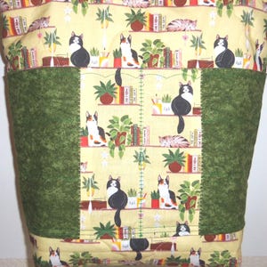 May include: A quilted fabric tote bag featuring a yellow background with a repeating pattern of cats, potted plants, and books on shelves. The bag includes two solid dark green vertical panels on the front, stitched with visible green thread.