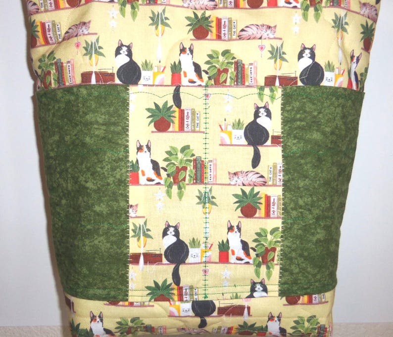 May include: A quilted fabric tote bag featuring a yellow background with a repeating pattern of cats, potted plants, and books on shelves. The bag includes two solid dark green vertical panels on the front, stitched with visible green thread.