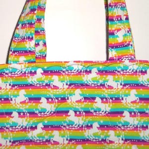 May include: A tote bag featuring a repeating pattern of white unicorns surrounded by stars on a background of horizontal rainbow stripes. The bag has two matching fabric handles and a scalloped top edge with visible quilted stitching.