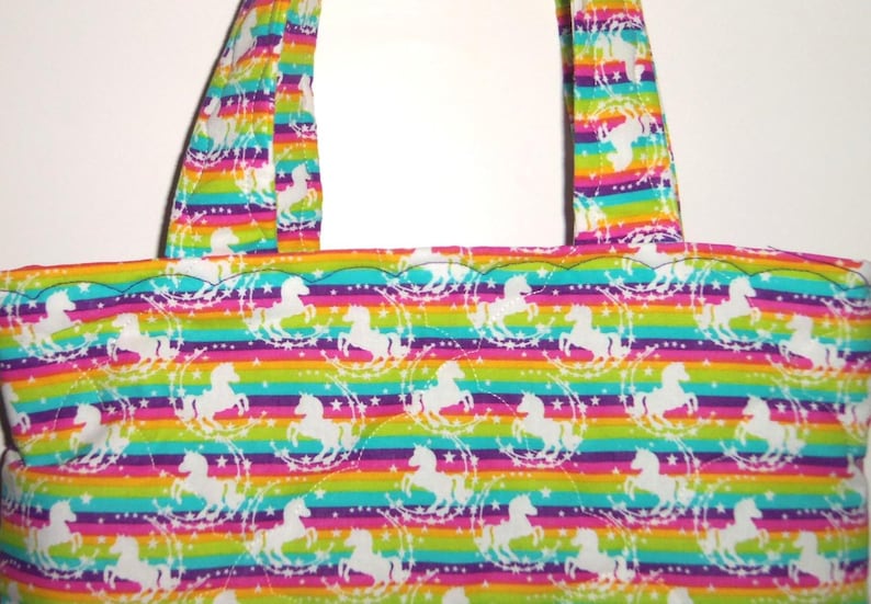 May include: A tote bag featuring a repeating pattern of white unicorns surrounded by stars on a background of horizontal rainbow stripes. The bag has two matching fabric handles and a scalloped top edge with visible quilted stitching.