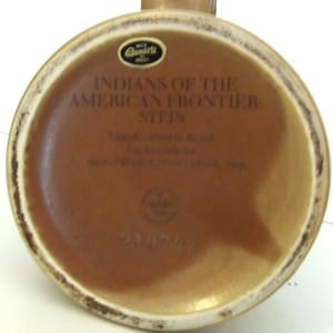 May include: The underside of a brown ceramic stein featuring a black sticker with the text Ceramarte Brazil. Printed text reads INDIANS OF THE AMERICAN FRONTIER STEIN, Handcrafted in Brazil, Exclusively for AVON PRODUCTS INC, 1985, and the number 210743.