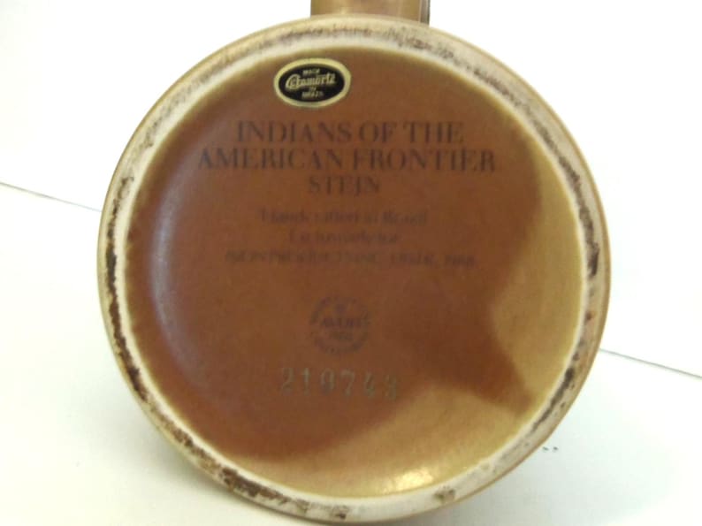 May include: The underside of a brown ceramic stein featuring a black sticker with the text Ceramarte Brazil. Printed text reads INDIANS OF THE AMERICAN FRONTIER STEIN, Handcrafted in Brazil, Exclusively for AVON PRODUCTS INC, 1985, and the number 210743.
