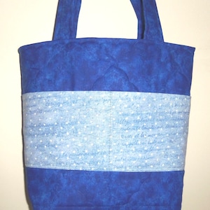 May include: A quilted tote bag featuring a deep blue mottled fabric on the top and bottom sections. A central horizontal band of light blue fabric displays a white script pattern with small white dots, finished with two fabric shoulder straps.