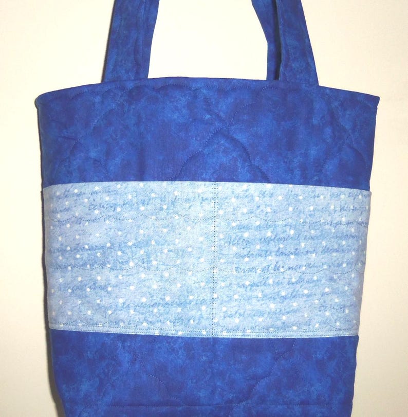 May include: A quilted tote bag featuring a deep blue mottled fabric on the top and bottom sections. A central horizontal band of light blue fabric displays a white script pattern with small white dots, finished with two fabric shoulder straps.