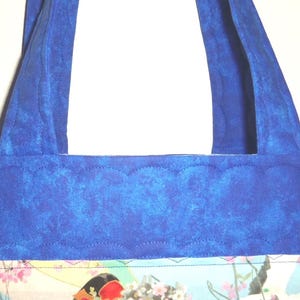 May include: A quilted tote bag featuring a vibrant blue fabric with a scalloped stitching pattern. The bag has two matching blue fabric handles and a rectangular central panel displaying a colorful graphic print with floral and abstract elements.