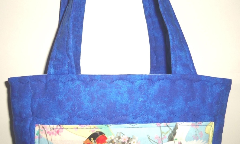 May include: A quilted tote bag featuring a vibrant blue fabric with a scalloped stitching pattern. The bag has two matching blue fabric handles and a rectangular central panel displaying a colorful graphic print with floral and abstract elements.
