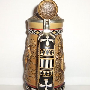May include: A tan ceramic beer stein featuring a metal lid with a textured circular top. The handle is black and white with geometric patterns and a cross design. The body displays embossed scenes of figures and structures with a decorative base.