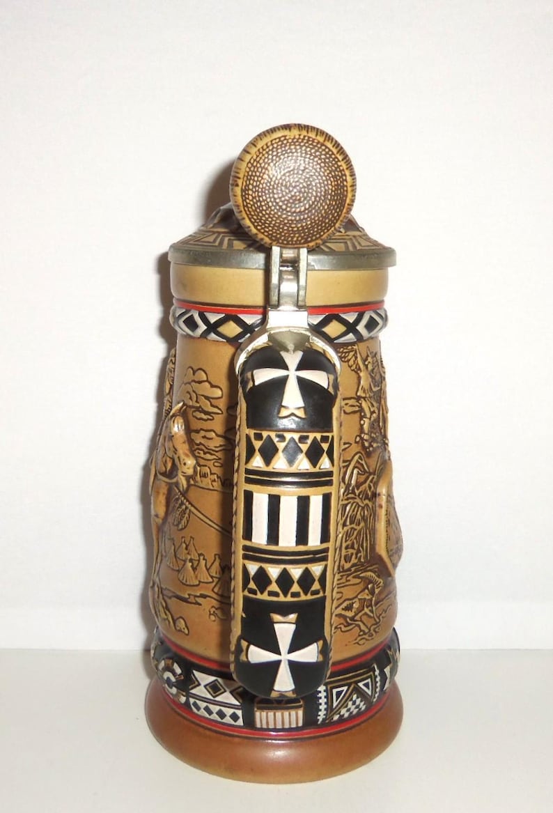 May include: A tan ceramic beer stein featuring a metal lid with a textured circular top. The handle is black and white with geometric patterns and a cross design. The body displays embossed scenes of figures and structures with a decorative base.