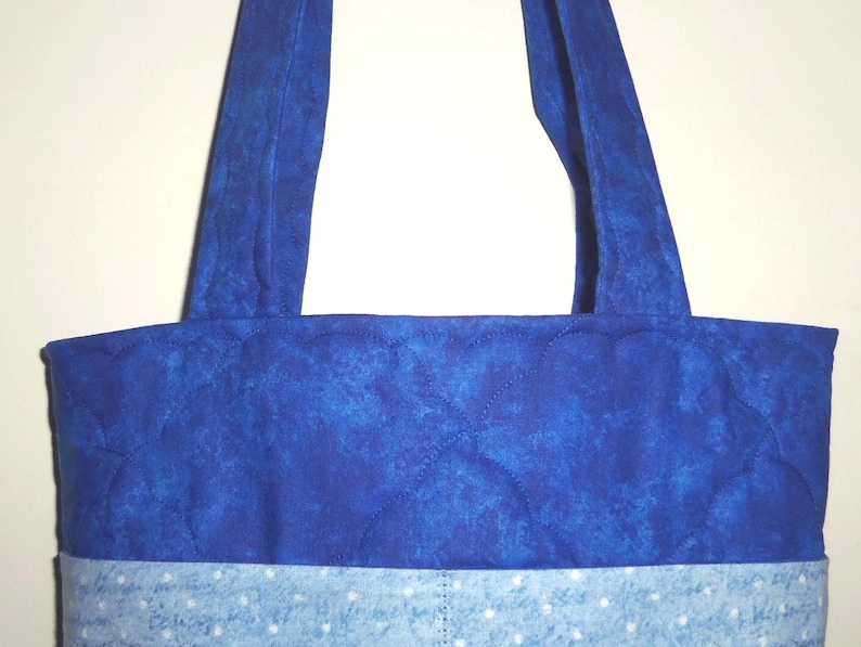 May include: A quilted tote bag featuring a deep royal blue upper section with a mottled texture and a lighter sky blue lower section adorned with a white polka dot pattern. The bag includes two matching royal blue fabric shoulder straps.
