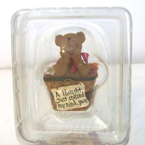 May include: A brown teddy bear figurine sits in a miniature basket with autumn leaves and a red bow. The basket has a sign that reads "A thought just crossed my mind...you". The figurine is encased in a clear plastic container.