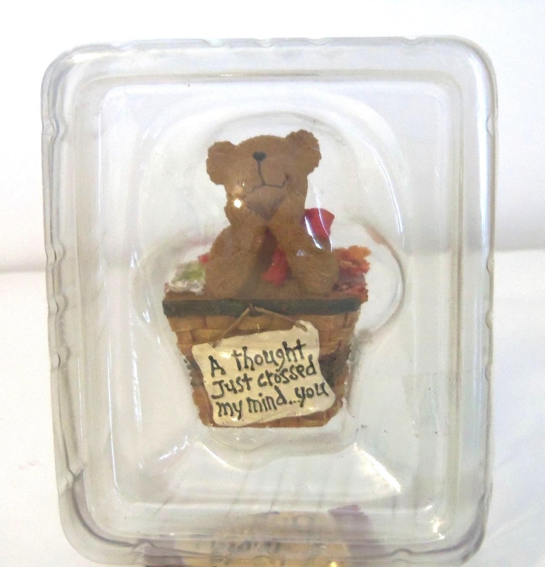 May include: A brown teddy bear figurine sits in a miniature basket with autumn leaves and a red bow. The basket has a sign that reads "A thought just crossed my mind...you". The figurine is encased in a clear plastic container.