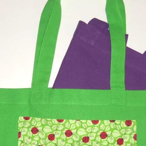 K&ouml;nnte beinhalten: Eine leuchtend gr&uuml;ne Tragetasche mit einer rechteckigen Vordertasche, die mit einem Muster aus roten Marienk&auml;fern und gr&uuml;nen Bl&auml;ttern bedruckt ist. Eine zweite, kleinere violette Tasche steckt in der gr&uuml;nen Tasche, deren Griffe oben herausragen.