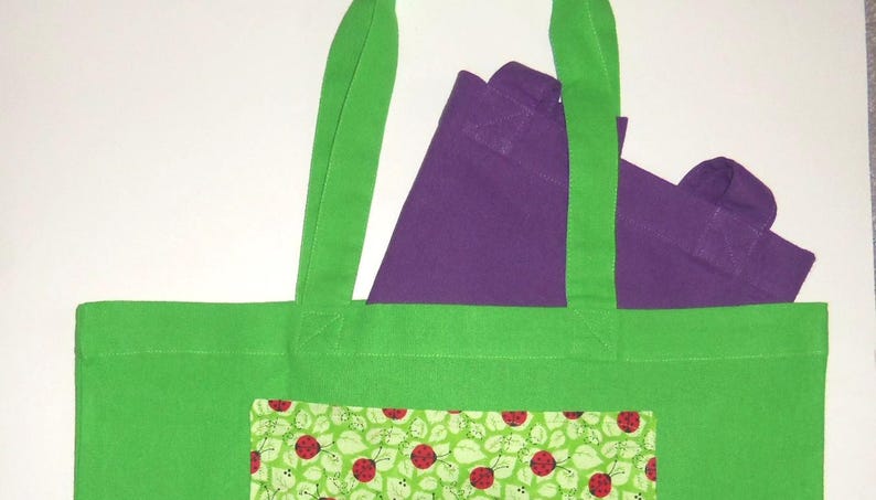 K&ouml;nnte beinhalten: Eine leuchtend gr&uuml;ne Tragetasche mit einer rechteckigen Vordertasche, die mit einem Muster aus roten Marienk&auml;fern und gr&uuml;nen Bl&auml;ttern bedruckt ist. Eine zweite, kleinere violette Tasche steckt in der gr&uuml;nen Tasche, deren Griffe oben herausragen.