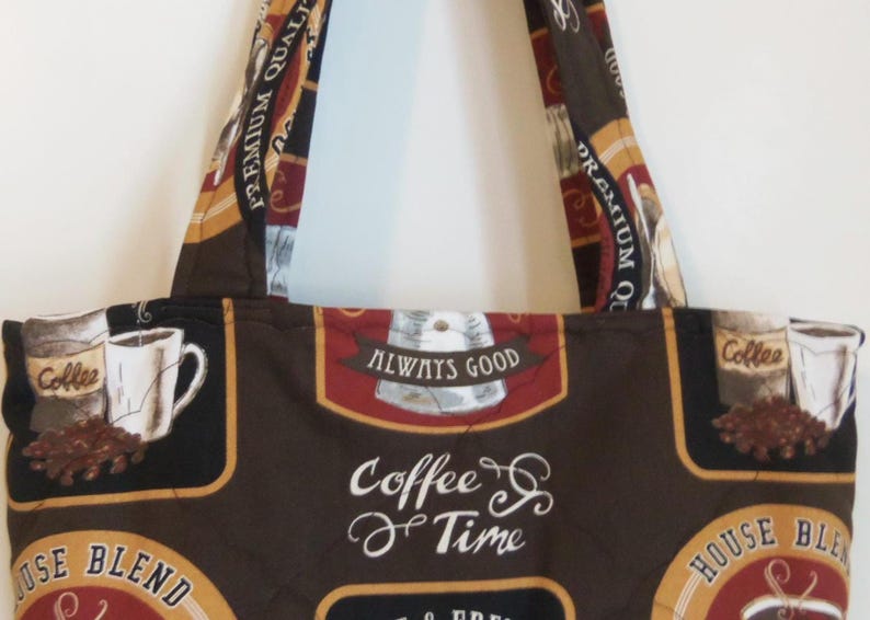 Puede incluir: Un bolso de tela con un estampado de caf&eacute; en tonos marr&oacute;n, negro y crema. El dise&ntilde;o incluye ilustraciones de tazas de caf&eacute;, granos y textos que dicen "Premium Quality", "Always Good", "Coffee Time" y "House Blend" por toda la superficie.