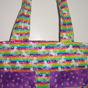 May include: A tote bag featuring a horizontal rainbow striped pattern with white silhouettes of horses surrounded by stars. The bag includes two matching fabric handles and a solid purple bottom section decorated with a subtle heart print pattern.