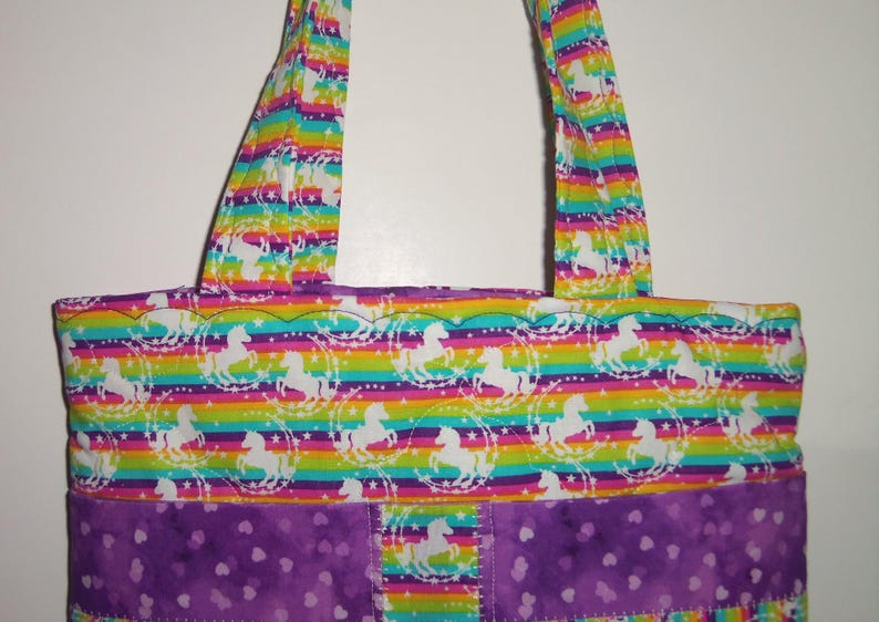 May include: A tote bag featuring a horizontal rainbow striped pattern with white silhouettes of horses surrounded by stars. The bag includes two matching fabric handles and a solid purple bottom section decorated with a subtle heart print pattern.