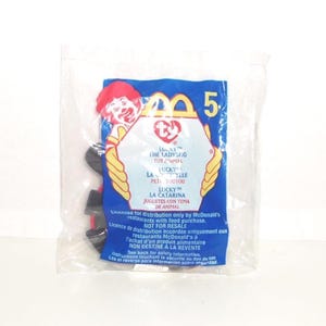 May include: A sealed clear plastic bag containing a McDonald's Ty Lucky the Ladybug toy. The packaging features a red Ronald McDonald graphic, the Ty logo, and text in English, French, and Spanish regarding distribution and safety information.