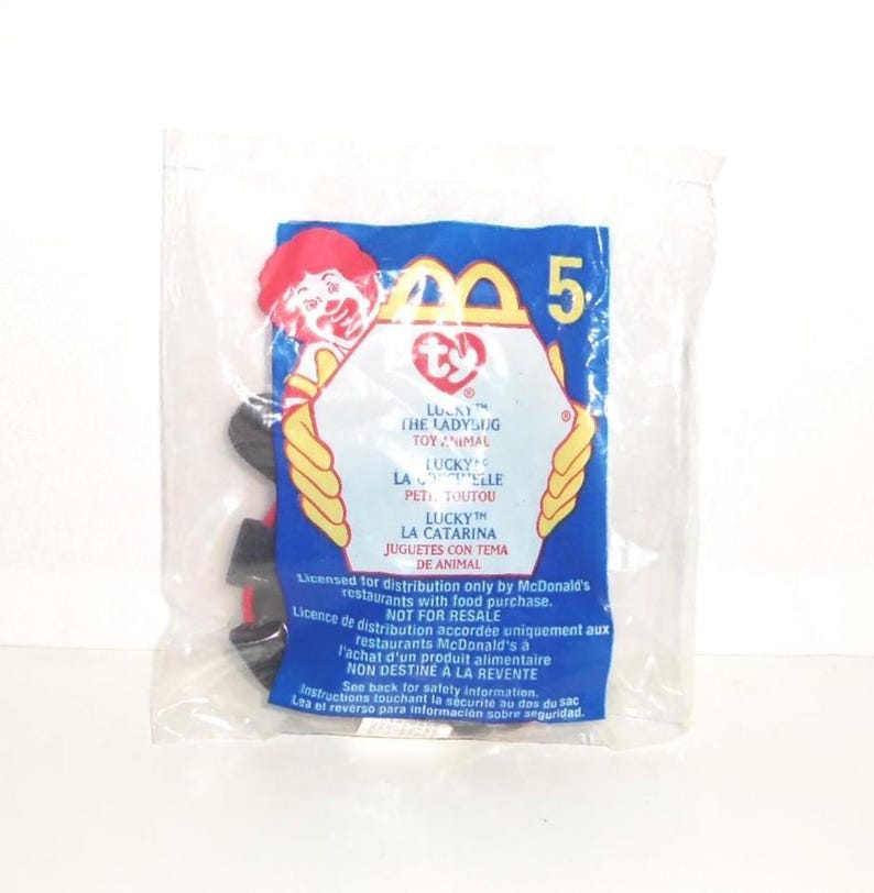 May include: A sealed clear plastic bag containing a McDonald's Ty Lucky the Ladybug toy. The packaging features a red Ronald McDonald graphic, the Ty logo, and text in English, French, and Spanish regarding distribution and safety information.