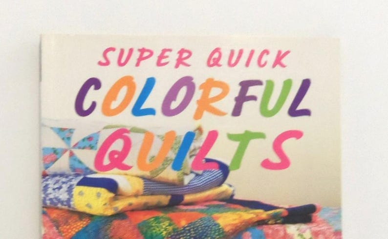 May include: A book titled SUPER QUICK COLORFUL QUILTS with a cover featuring a stack of folded fabric quilts in various patterns and bright colors including blue, yellow, red, and green. The text is printed in large, multi-colored sans-serif letters.
