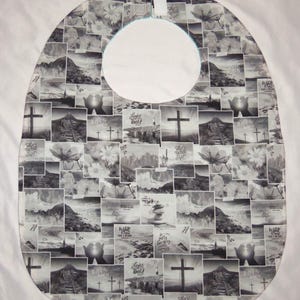 May include: A bib featuring a black and white collage print of various religious and nature-themed images, including crosses, mountains, flowers, and stacked stones. The fabric has a rounded shape with a central neck opening and a hook-and-loop closure.