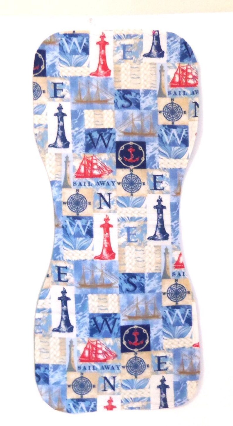 May include: A nautical-themed baby burp cloth with a patchwork design. The cloth features blue and white squares with images of lighthouses, sailboats, compasses, and the words "SAIL AWAY". The cloth is shaped to fit over the shoulder.