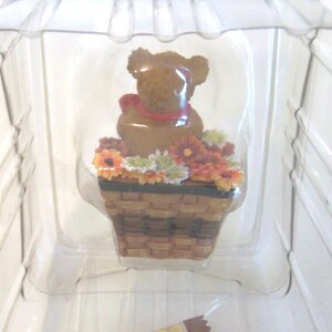 May include: A brown teddy bear figurine with a red bow, sitting in a brown basket filled with colorful flowers. The figurine is encased in a clear plastic container.