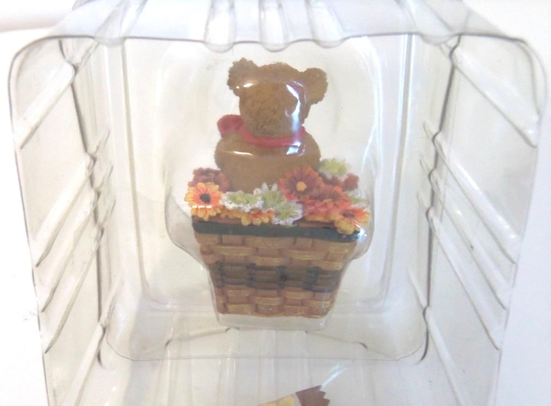May include: A brown teddy bear figurine with a red bow, sitting in a brown basket filled with colorful flowers. The figurine is encased in a clear plastic container.
