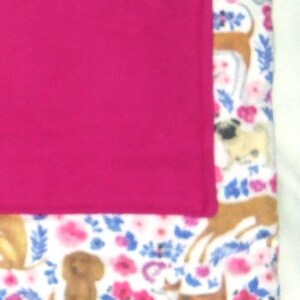 May include: A white fleece blanket with a pink pocket and a repeating pattern of dogs and flowers. The dogs are brown, black, and white.