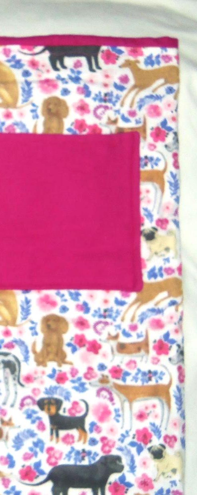 May include: A white fleece blanket with a pink pocket and a repeating pattern of dogs and flowers. The dogs are brown, black, and white.