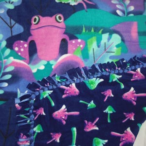 May include: A fleece blanket with a fantasy design. The blanket displays a pink frog in a colourful forest scene with purple, teal, and green elements. The lower section has a pattern of pink and green mushrooms on a navy blue background. The edges are finished with a navy blue fringe.