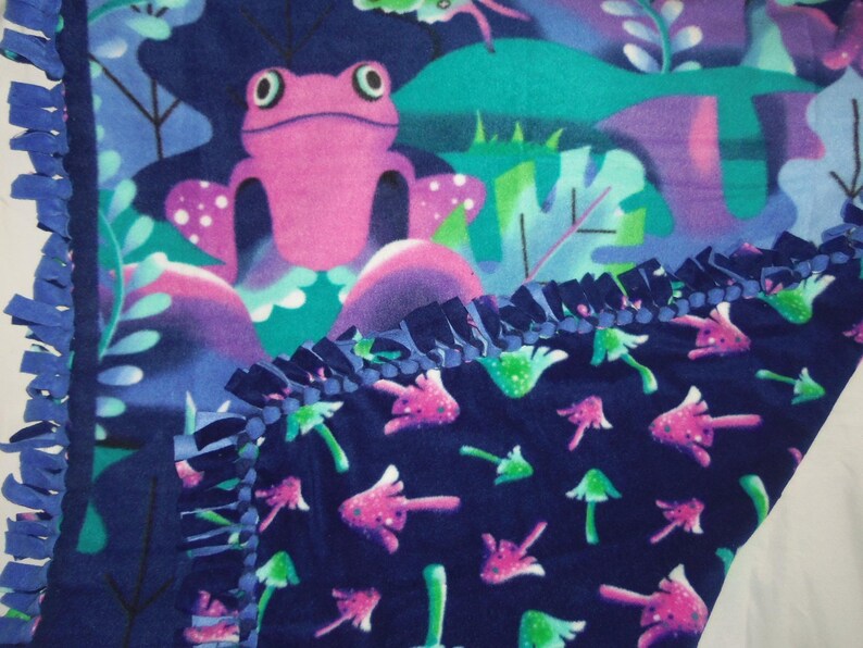 May include: A fleece blanket with a fantasy design. The blanket displays a pink frog in a colourful forest scene with purple, teal, and green elements. The lower section has a pattern of pink and green mushrooms on a navy blue background. The edges are finished with a navy blue fringe.