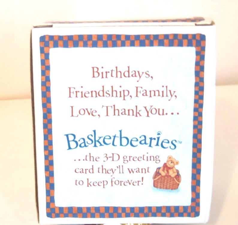 Op de afbeelding: Een witte doos met een blauw en oranje geruite rand. De tekst luidt: "Verjaardagen, Vriendschap, Familie, Liefde, Dank u... Basketbearies...de 3D-wenskaart die ze voor altijd willen bewaren!" Er is ook een kleine illustratie van een teddybeer in een mand.