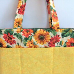 May include: A yellow quilted tote bag with a sunflower print. The bag has a large pocket on the front and two handles.