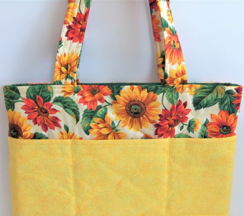 May include: A yellow quilted tote bag with a sunflower print. The bag has a large pocket on the front and two handles.