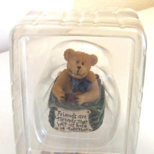 May include: A small, tan teddy bear figurine with a blue bow tie, seated in a green basket. The figurine is inside a clear plastic container. A sign on the basket reads "Friends are threads that help us hold it together."