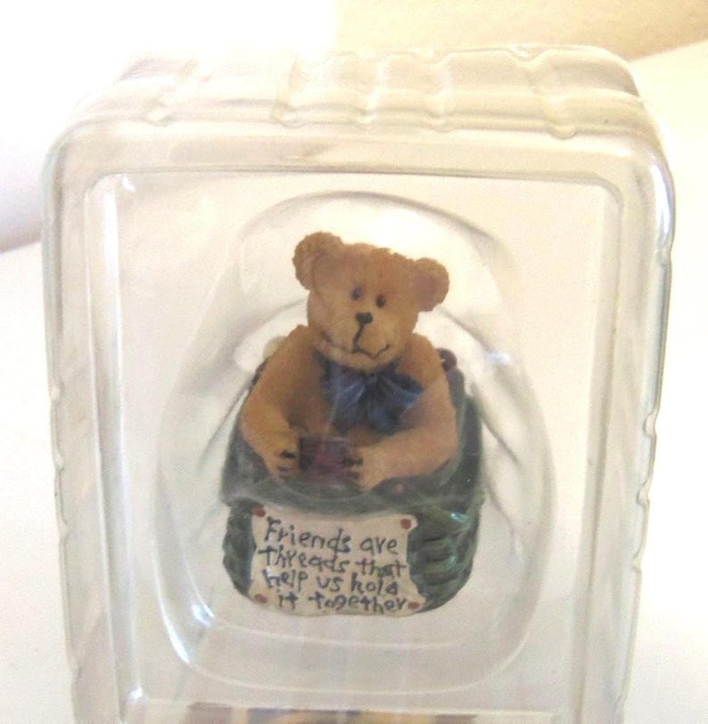 May include: A small, tan teddy bear figurine with a blue bow tie, seated in a green basket. The figurine is inside a clear plastic container. A sign on the basket reads "Friends are threads that help us hold it together."