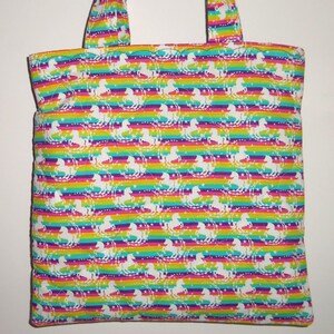 May include: A fabric tote bag featuring a repeating pattern of white unicorns and stars set against a vibrant horizontal rainbow striped background. The bag includes two matching fabric handles at the top and has a quilted texture throughout.