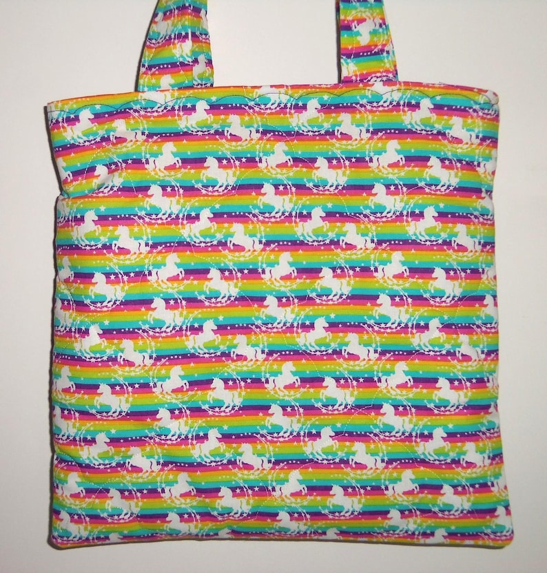 May include: A fabric tote bag featuring a repeating pattern of white unicorns and stars set against a vibrant horizontal rainbow striped background. The bag includes two matching fabric handles at the top and has a quilted texture throughout.