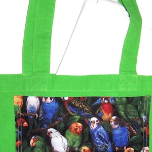 May include: A bright green tote bag featuring a rectangular central panel printed with a detailed pattern of various colorful parrots and parakeets perched among dark green foliage. The bag has two matching green fabric shoulder straps.