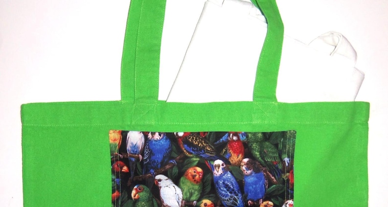 May include: A bright green tote bag featuring a rectangular central panel printed with a detailed pattern of various colorful parrots and parakeets perched among dark green foliage. The bag has two matching green fabric shoulder straps.