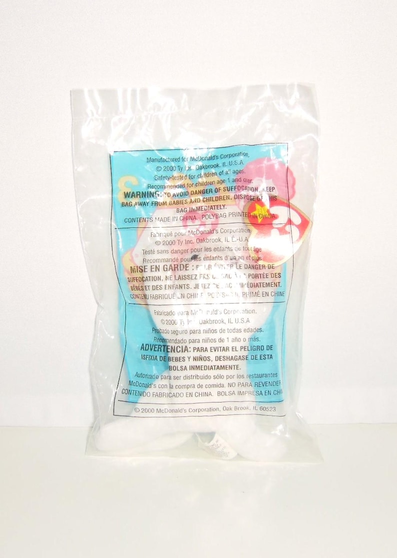 May include: A plush toy sealed in a clear plastic bag with a light blue label featuring text in English, French, and Spanish. The label includes the Ty logo, McDonald's Corporation branding, and safety warnings dated 2000. The toy is partially visible inside the packaging, showing white fabric and a red heart-shaped tag.