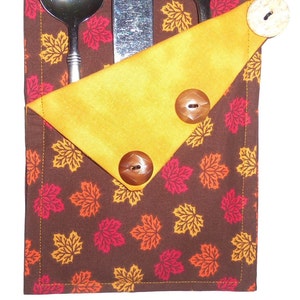 May include: A brown fabric cutlery holder with a yellow fabric flap. The holder is decorated with a pattern of orange, red, and yellow maple leaves. The flap has three brown buttons.