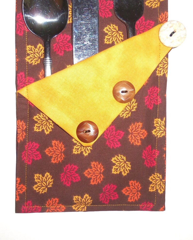 May include: A brown fabric cutlery holder with a yellow fabric flap. The holder is decorated with a pattern of orange, red, and yellow maple leaves. The flap has three brown buttons.