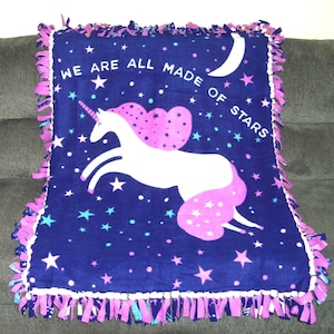 Könnte beinhalten: Eine marineblaue Fleece-Decke mit einem weißen Einhorn-Design und dem Text "WE ARE ALL MADE OF STARS". Die Decke hat einen rosa-violetten Fransenrand und ist mit weißen, blauen und rosa Sternen bedeckt.