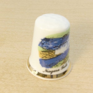 May include: White ceramic thimble with a gold rim and a colorful image of Niagara Falls. The thimble has the words "Niagara Falls" printed on the bottom.