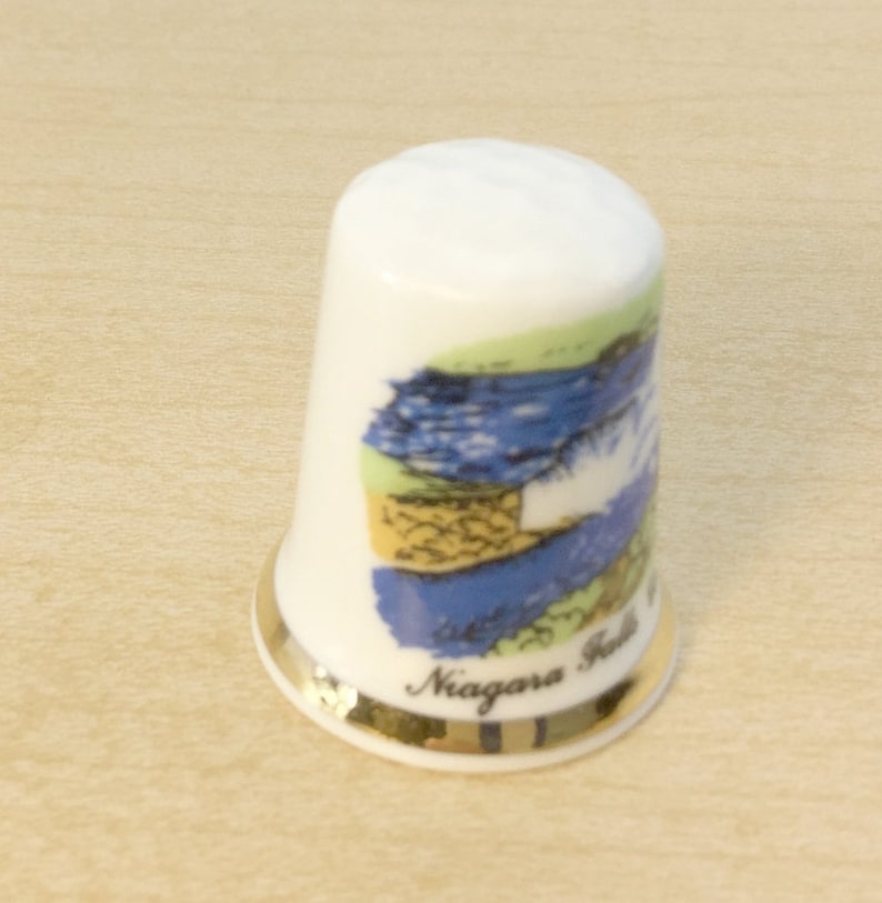 May include: White ceramic thimble with a gold rim and a colorful image of Niagara Falls. The thimble has the words "Niagara Falls" printed on the bottom.