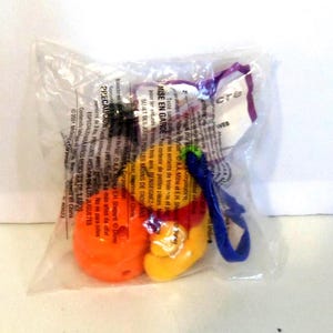 K&ouml;nnte beinhalten: Ein kleiner, versiegelter, durchsichtiger Plastikbeutel mit einem bunten Spielzeug in Orange, Gelb und Rot. An der Seite befindet sich eine blaue Kunststoffschlaufe. Die Verpackung ist mit Text in mehreren Sprachen bedruckt, darunter "MISE EN GARDE" und "2001 McDonald's Corporation".