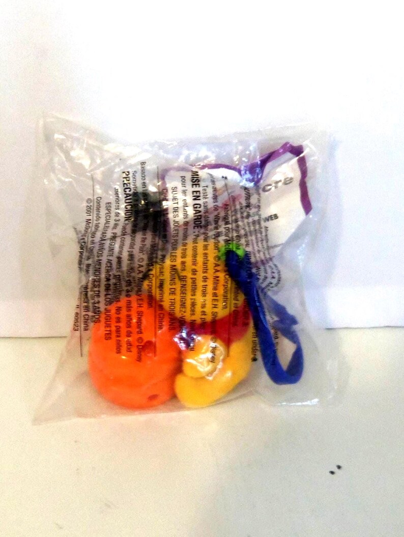 K&ouml;nnte beinhalten: Ein kleiner, versiegelter, durchsichtiger Plastikbeutel mit einem bunten Spielzeug in Orange, Gelb und Rot. An der Seite befindet sich eine blaue Kunststoffschlaufe. Die Verpackung ist mit Text in mehreren Sprachen bedruckt, darunter "MISE EN GARDE" und "2001 McDonald's Corporation".