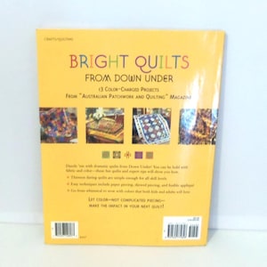May include: A yellow softcover book titled Bright Quilts from Down Under, featuring 13 color-charged projects from Australian Patchwork and Quilting magazine. The back cover displays four small quilt photos, text descriptions, and a barcode.